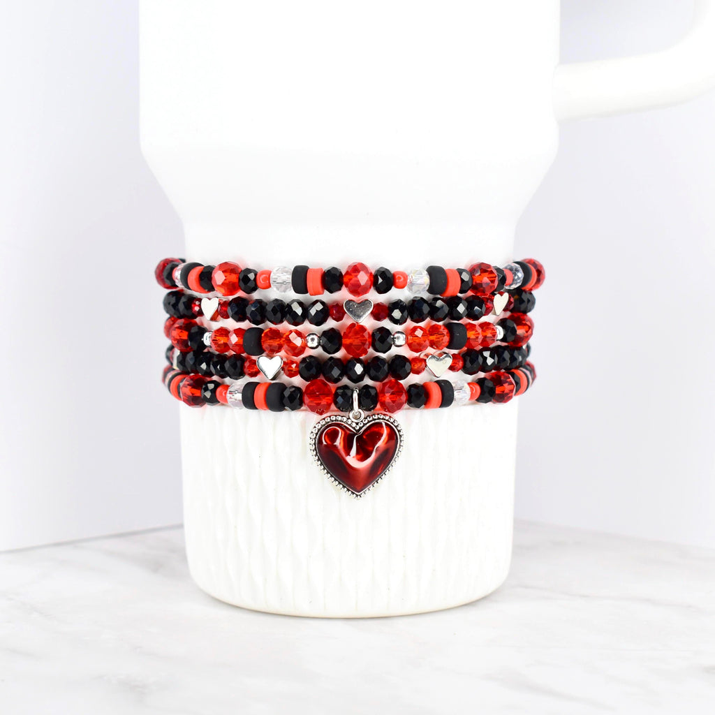 A stack of beaded bracelets in red, black, and silver, wrapped around a white Stanley tumbler. The bracelets feature various bead sizes and shapes, with a heart-shaped charm dangling from the bottom. The beads are arranged in a repeating pattern.