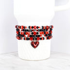 A stack of beaded bracelets in red, black, and silver, wrapped around a white Stanley tumbler. The bracelets feature various bead sizes and shapes, with a heart-shaped charm dangling from the bottom. The beads are arranged in a repeating pattern.