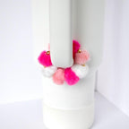 A white tumbler with a handle, adorned with a colorful pom-pom accessory. The pom-poms are in shades of pink and white, with gold accents. The tumbler is set against a white background.