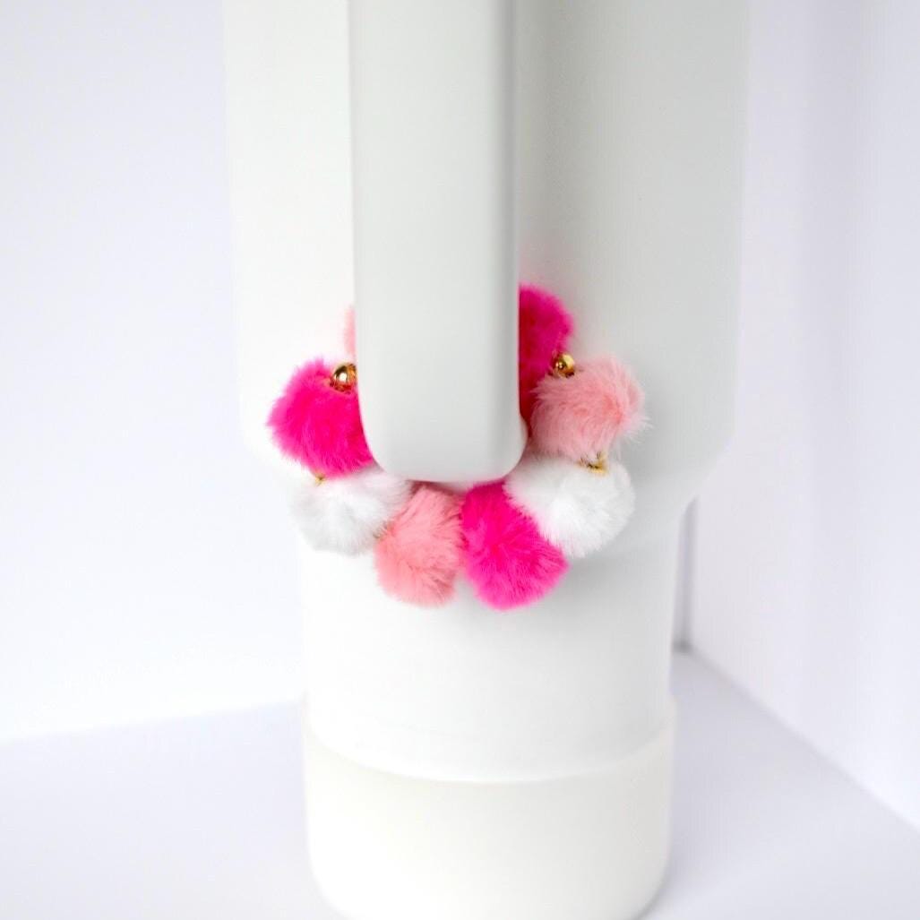 A white tumbler with a handle, adorned with a colorful pom-pom accessory. The pom-poms are in shades of pink and white, with gold accents. The tumbler is set against a white background.