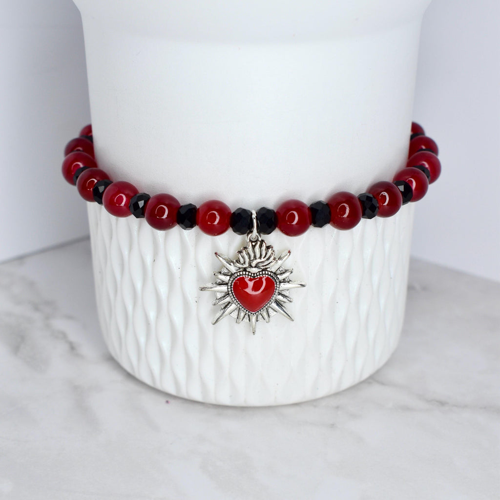 A red beaded bracelet with black accents and a silver charm. The charm is a stylized heart with a flame design. The bracelet is displayed on a white ceramic container.