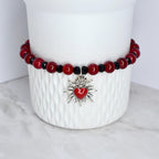 A red beaded bracelet with black accents and a silver charm. The charm is a stylized heart with a flame design. The bracelet is displayed on a white ceramic container.