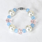 A tumbler handle bracelet featuring large, white faux pearls, light blue beads, and faceted pink crystal beads. The bracelet is connected with a silver-colored magnetic clasp. The beads are strung together to form a circular shape.