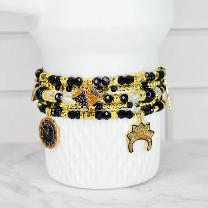 A stack of beaded bracelets in black, gold, and clear beads with gold charms. The charms include a coin, a crown, and a geometric design. The bracelets are displayed on a white, textured vase.