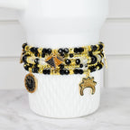 A stack of beaded bracelets in black, gold, and clear beads with gold charms. The charms include a coin, a crown, and a geometric design. The bracelets are displayed on a white, textured vase.