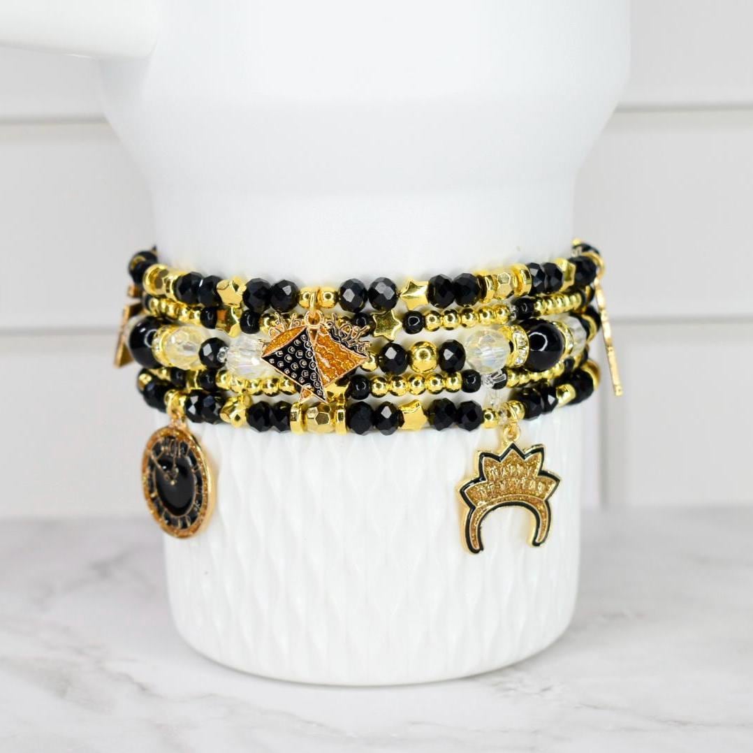 A stack of beaded bracelets in black, gold, and clear beads with gold charms. The charms include a coin, a crown, and a geometric design. The bracelets are displayed on a white, textured vase.