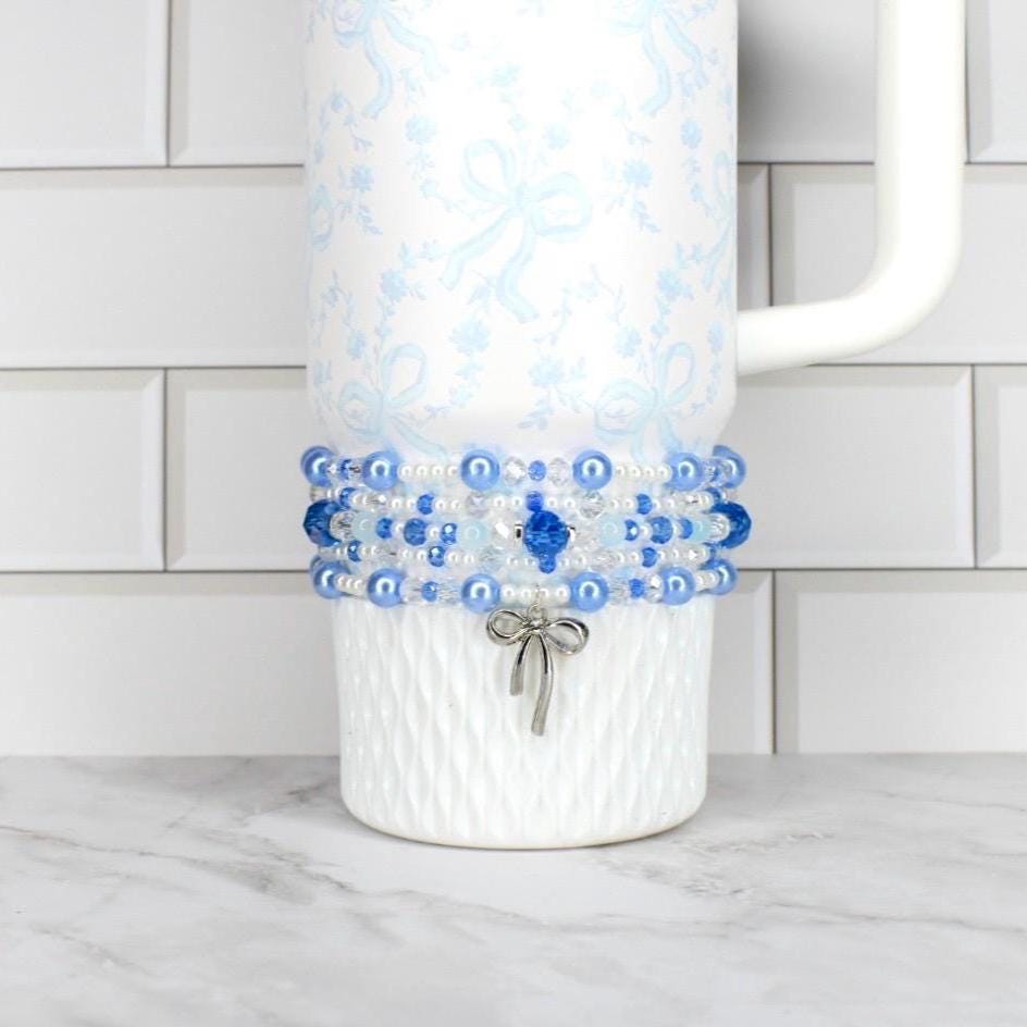 A white tumbler with a blue floral pattern and a beaded wrap. The wrap features blue and white beads, with a silver bow charm. The tumbler has a white handle and is on a white surface.