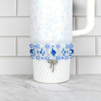 A white tumbler with a blue floral pattern and a beaded wrap. The wrap features blue and white beads, with a silver bow charm. The tumbler has a white handle and is on a white surface.