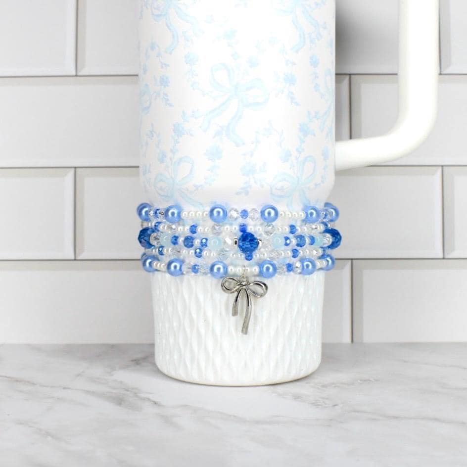 A white tumbler with a blue floral pattern and a beaded wrap. The wrap features blue and white beads, with a silver bow charm. The tumbler has a white handle and is on a white surface.