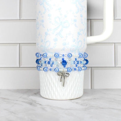 A white tumbler with a blue floral pattern and a beaded wrap. The wrap features blue and white beads, with a silver bow charm. The tumbler has a white handle and is on a white surface.