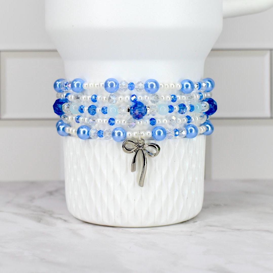 A Stanley tumbler with a textured surface is adorned with a beaded bracelet. The bracelet features layers of blue and white beads, with a silver bow charm hanging from the bottom. The beads are various sizes and shapes.