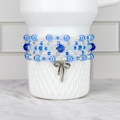 A Stanley tumbler with a textured surface is adorned with a beaded bracelet. The bracelet features layers of blue and white beads, with a silver bow charm hanging from the bottom. The beads are various sizes and shapes.