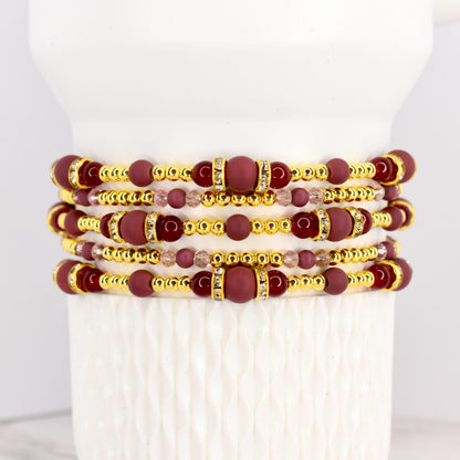 Stack of beaded bracelets in shades of burgundy, gold, and clear crystals. The bracelets feature a mix of round and cylindrical beads. The gold beads have a metallic finish, and the burgundy beads have a matte finish.