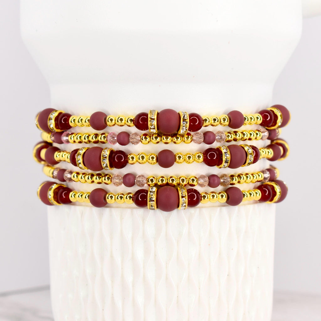 Stack of beaded bracelets in shades of burgundy, gold, and clear crystals. The bracelets feature a mix of round and cylindrical beads. The gold beads have a metallic finish, and the burgundy beads have a matte finish.