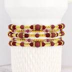 Stack of beaded bracelets in shades of burgundy, gold, and clear crystals. The bracelets feature a mix of round and cylindrical beads. The gold beads have a metallic finish, and the burgundy beads have a matte finish.