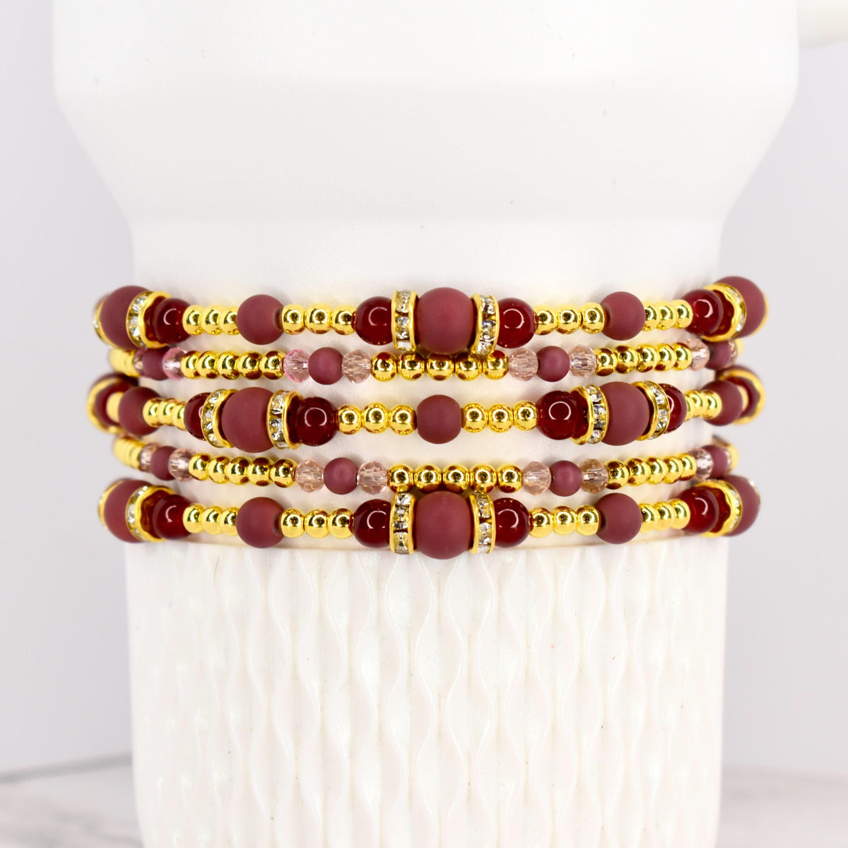 Stack of beaded bracelets in shades of burgundy, gold, and clear crystals. The bracelets feature a mix of round and cylindrical beads. The gold beads have a metallic finish, and the burgundy beads have a matte finish.