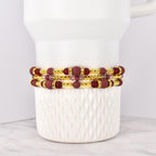 Three beaded bracelets with burgundy and gold accents. The bracelets feature burgundy matte beads, gold-colored spacer beads, and decorative gold-colored bands with clear rhinestones. The bracelets are stacked on a white ceramic container.
