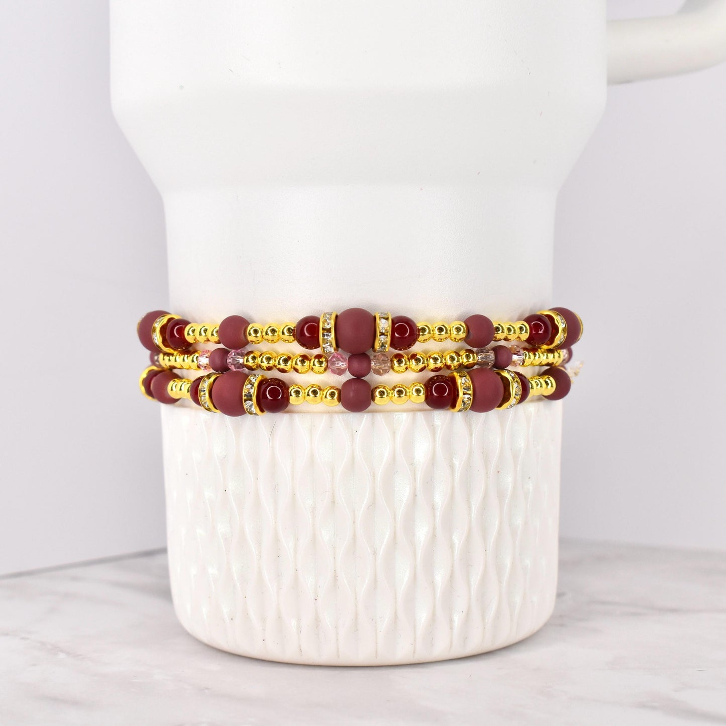 Three beaded bracelets with burgundy and gold accents. The bracelets feature burgundy matte beads, gold-colored spacer beads, and decorative gold-colored bands with clear rhinestones. The bracelets are stacked on a white ceramic container.