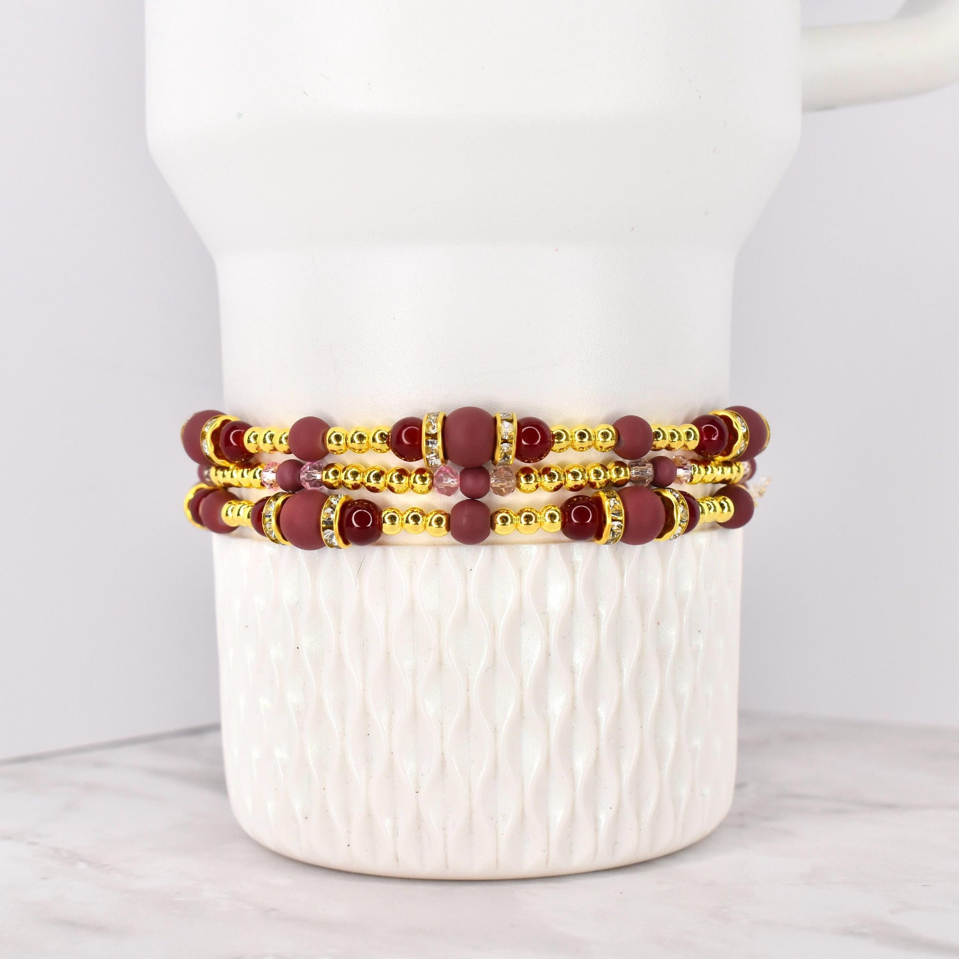 Three beaded bracelets with burgundy and gold accents. The bracelets feature burgundy matte beads, gold-colored spacer beads, and decorative gold-colored bands with clear rhinestones. The bracelets are stacked on a white ceramic container.