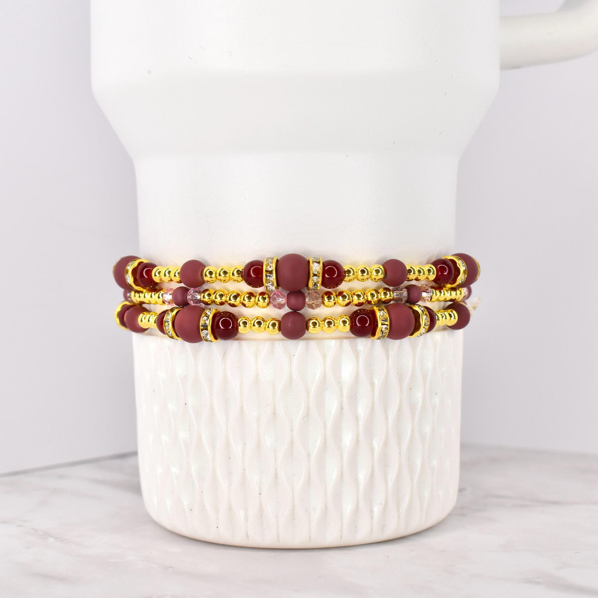 Three beaded bracelets with burgundy and gold accents. The bracelets feature burgundy matte beads, gold-colored spacer beads, and decorative gold-colored bands with clear rhinestones. The bracelets are stacked on a white ceramic container.