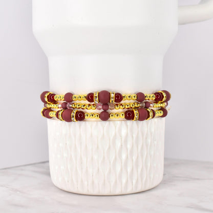 Three beaded bracelets with burgundy and gold accents. The bracelets feature burgundy matte beads, gold-colored spacer beads, and decorative gold-colored bands with clear rhinestones. The bracelets are stacked on a white ceramic container.