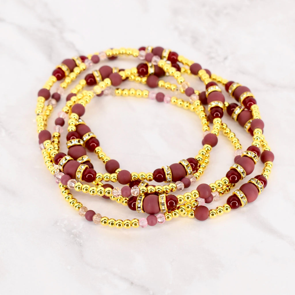 Gold-colored waist beads with burgundy and pink beads. The waist beads have a mix of small gold beads, larger burgundy beads, and decorative sections with rhinestones. The beads are arranged in multiple strands.