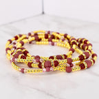 A collection of gold-colored beaded waist beads with burgundy and pink accents. The beads are arranged in multiple strands, with larger burgundy beads and smaller gold and pink beads. Some beads have rhinestone accents.