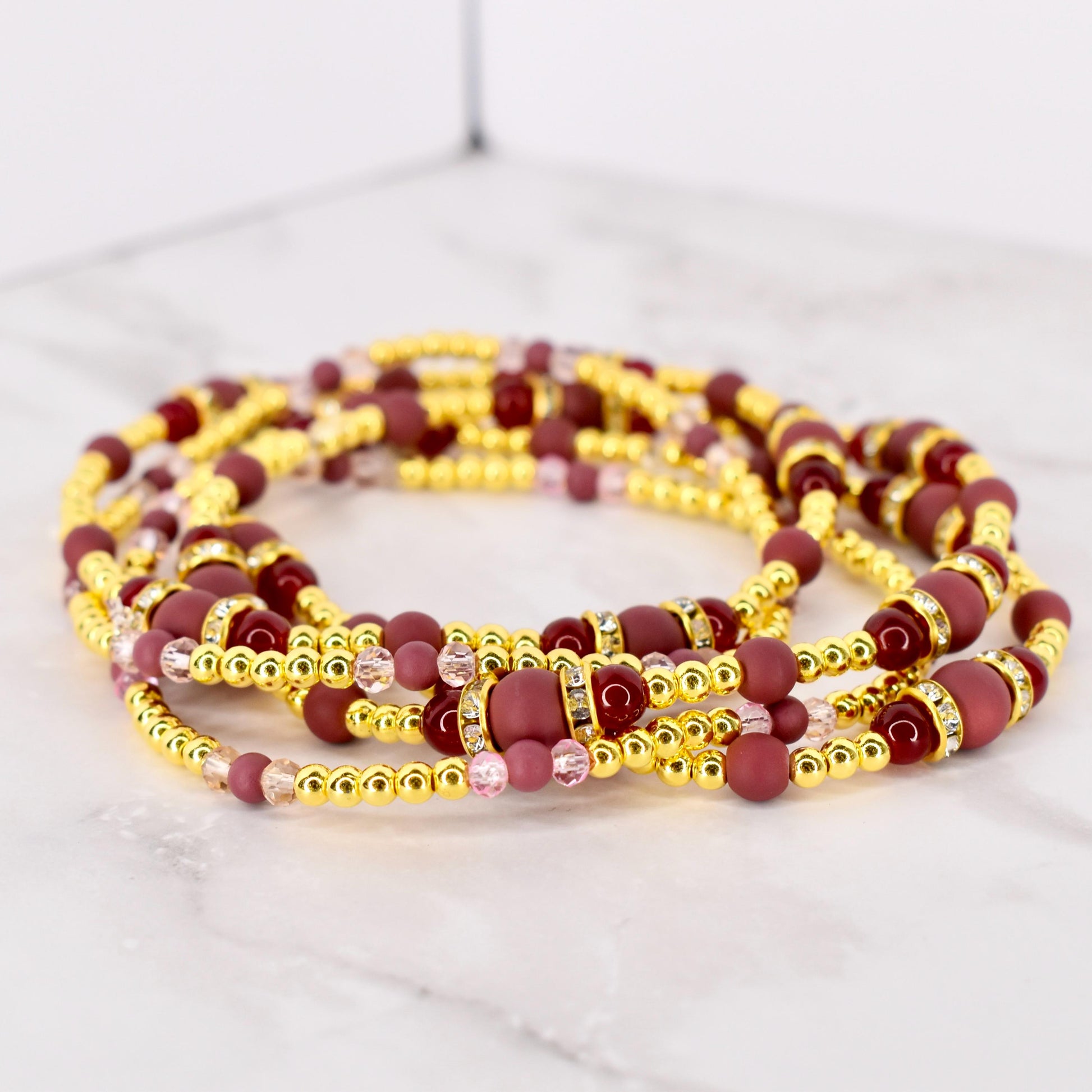 A collection of gold-colored beaded waist beads with burgundy and pink accents. The beads are arranged in multiple strands, with larger burgundy beads and smaller gold and pink beads. Some beads have rhinestone accents.