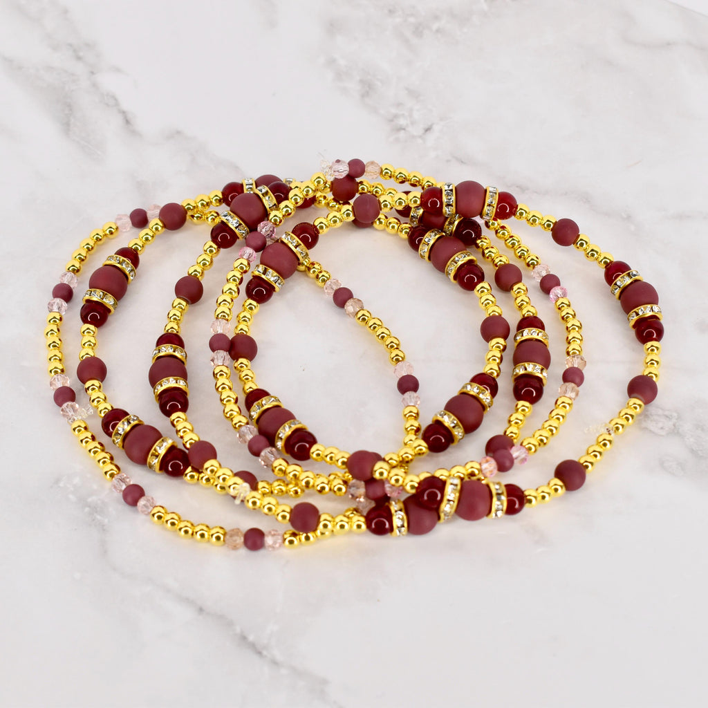 Strands of waist beads in a layered arrangement. The beads are a mix of gold, burgundy, and pink. The burgundy beads are cylindrical, while the gold beads are small and round. The pink beads are small and translucent.