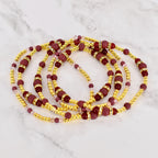 Strands of waist beads in a layered arrangement. The beads are a mix of gold, burgundy, and pink. The burgundy beads are cylindrical, while the gold beads are small and round. The pink beads are small and translucent.