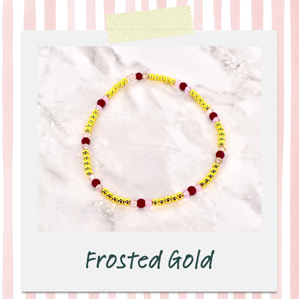 A beaded bracelet with gold beads, interspersed with burgundy and pink beads. The bracelet is displayed on a white surface, with the words "Frosted Gold" in a script font at the bottom.