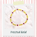 A beaded bracelet with gold beads, interspersed with burgundy and pink beads. The bracelet is displayed on a white surface, with the words "Frosted Gold" in a script font at the bottom.