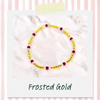 A beaded bracelet with gold beads, interspersed with burgundy and pink beads. The bracelet is displayed on a white surface, with the words "Frosted Gold" in a script font at the bottom.
