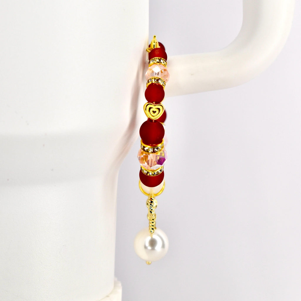 A gold-toned tumbler handle bracelet with red, pink, and clear beads, and a gold heart charm. The bracelet has a dangling white pearl at the bottom. The bracelet is displayed against a white background.