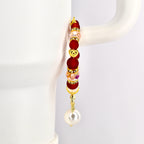 A gold-toned tumbler handle bracelet with red, pink, and clear beads, and a gold heart charm. The bracelet has a dangling white pearl at the bottom. The bracelet is displayed against a white background.