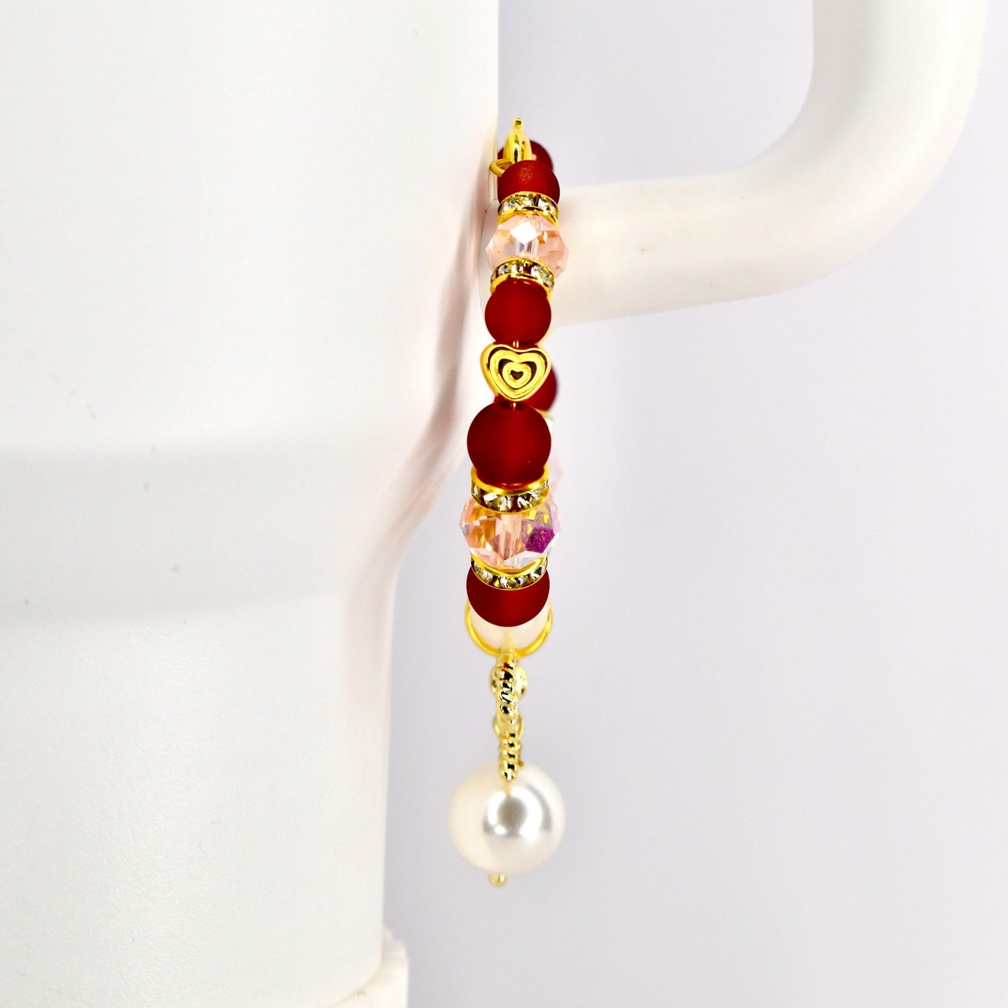 A gold-toned tumbler handle bracelet with red, pink, and clear beads, and a gold heart charm. The bracelet has a dangling white pearl at the bottom. The bracelet is displayed against a white background.