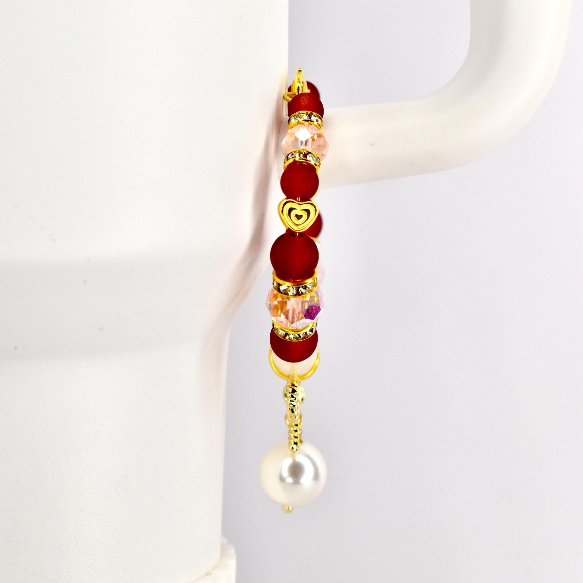 A gold-toned tumbler handle bracelet with red, pink, and clear beads, and a gold heart charm. The bracelet has a dangling white pearl at the bottom. The bracelet is displayed against a white background.