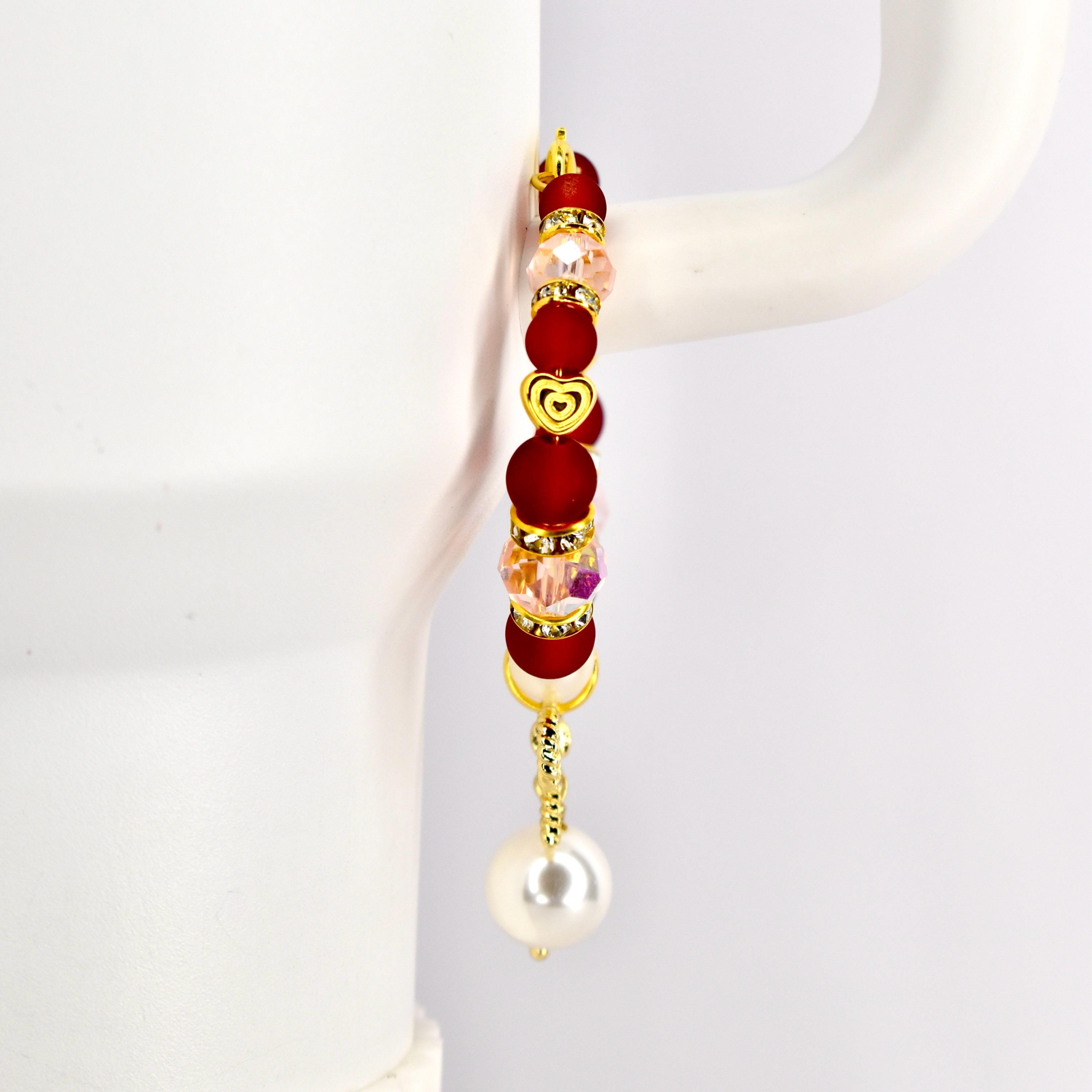 A gold-toned tumbler handle bracelet with red, pink, and clear beads, and a gold heart charm. The bracelet has a dangling white pearl at the bottom. The bracelet is displayed against a white background.