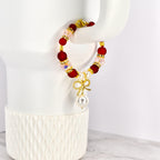 A beaded red velvet cake tumbler bracelet featuring red, pink, and gold beads. A gold bow charm with a large white pearl hangs from the bracelet. The bracelet is displayed on a white tumbler.
