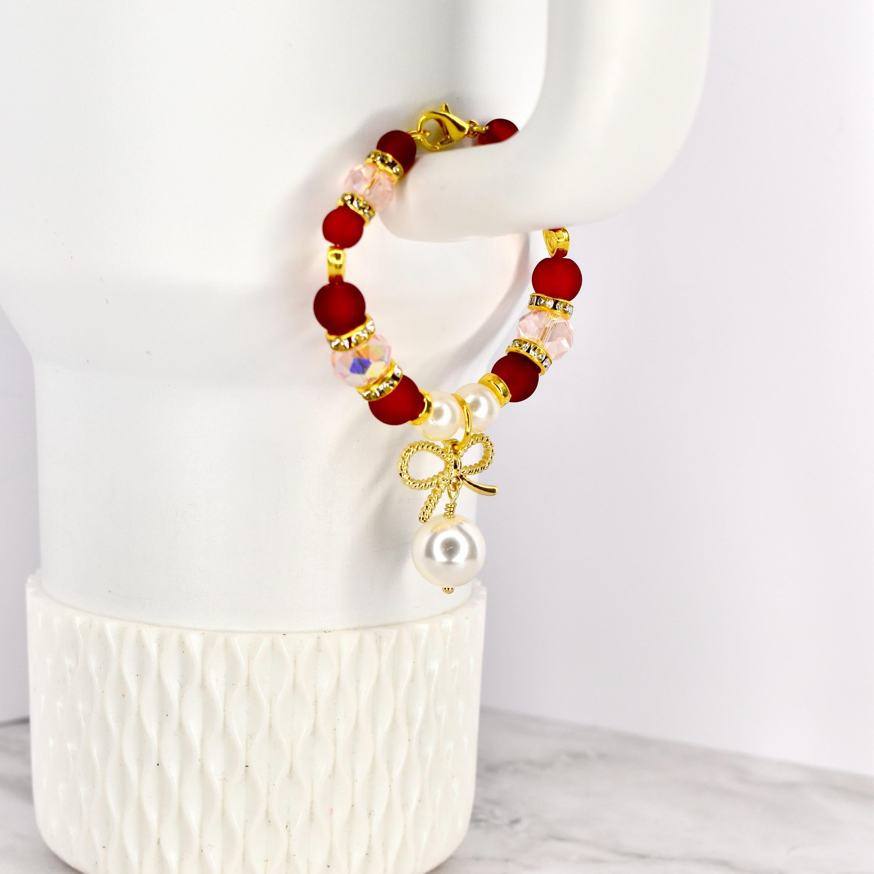 A beaded red velvet cake tumbler bracelet featuring red, pink, and gold beads. A gold bow charm with a large white pearl hangs from the bracelet. The bracelet is displayed on a white tumbler.
