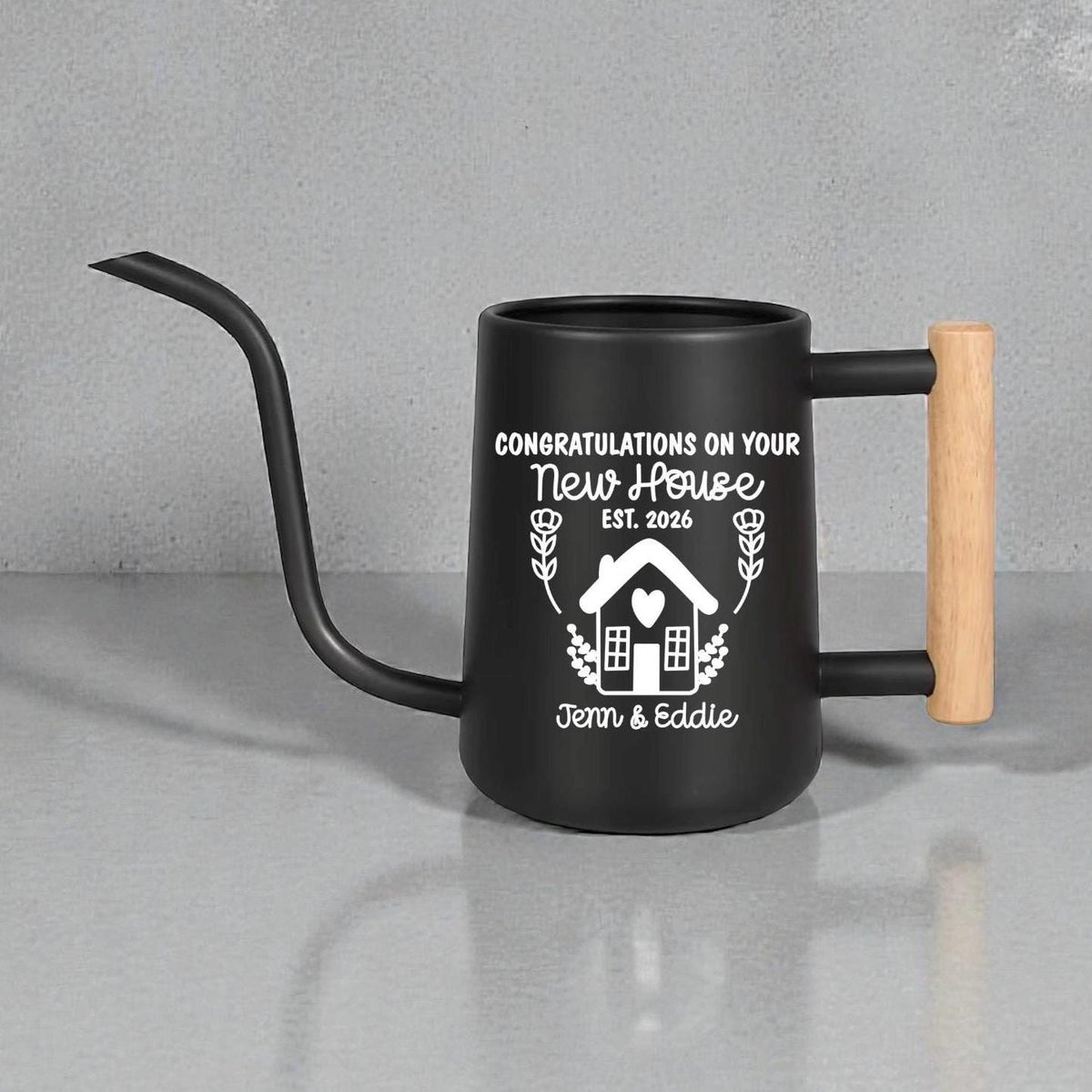 Black gooseneck watering can with wood handle. The design on the side is of a house with botanical accents in white vinyl and the words 'congratulations on your new house' above the design and the  names of the new home owners below the design.