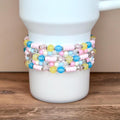 A white ceramic mug is adorned with multiple beaded bracelets. The bracelets feature a pastel color palette of pink, blue, yellow, and clear beads. The bracelets are wrapped around the mug.
