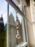 A decorative crystal suncatcher hanging from a silver chain. The suncatcher features a large, faceted crystal ball at the bottom, with a strand of iridescent and dark beads above. The background shows a window with a view of trees.