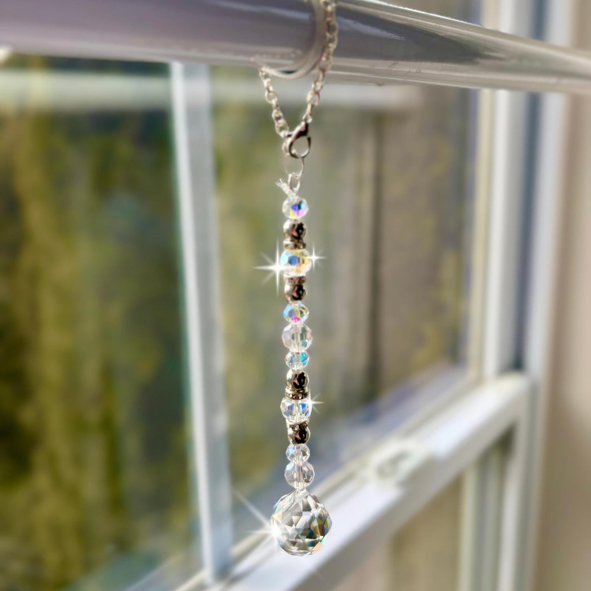 A decorative window sun catcher featuring a silver chain with a lobster clasp, clear and iridescent beads, and a faceted crystal ball at the bottom. The beads are interspersed with silver-colored spacers.