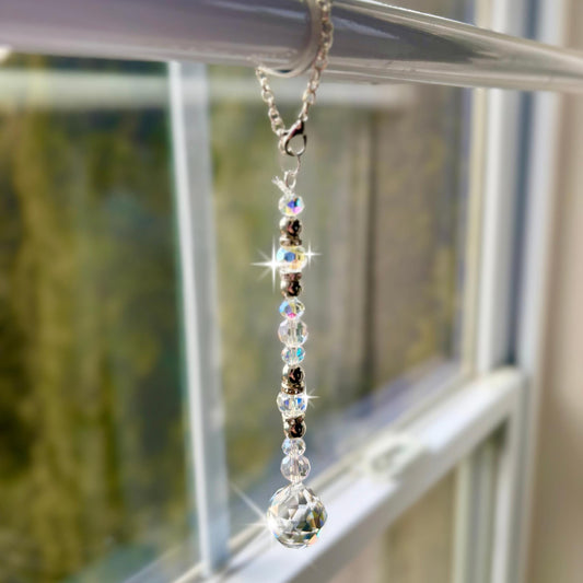 A decorative window sun catcher featuring a silver chain with a lobster clasp, clear and iridescent beads, and a faceted crystal ball at the bottom. The beads are interspersed with silver-colored spacers.