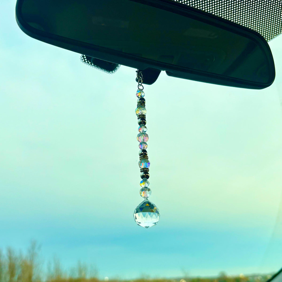 A car charm featuring a string of iridescent beads and a faceted crystal ball. The charm hangs from a rearview mirror, reflecting light. The background is a soft blue sky.