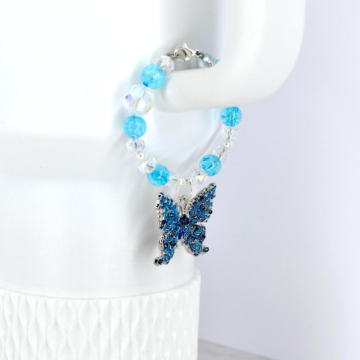 A blue beaded bracelet with a silver butterfly charm. The bracelet features light blue and clear beads. The butterfly charm is detailed with dark blue enamel and silver accents. The bracelet is displayed on a white surface.