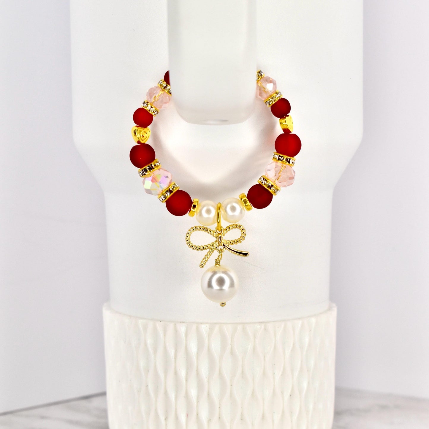 A beaded tumbler handle bracelet with red, pink, and gold accents. The bracelet features a gold bow charm with a dangling pearl. The bracelet is displayed on a white Stanley.