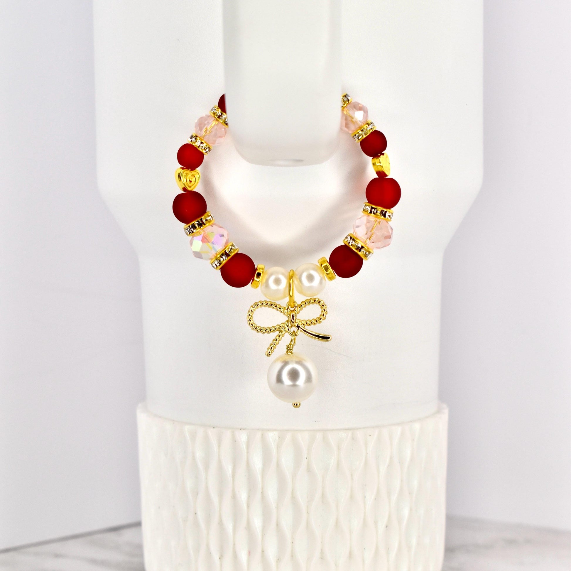 A beaded tumbler handle bracelet with red, pink, and gold accents. The bracelet features a gold bow charm with a dangling pearl. The bracelet is displayed on a white Stanley.
