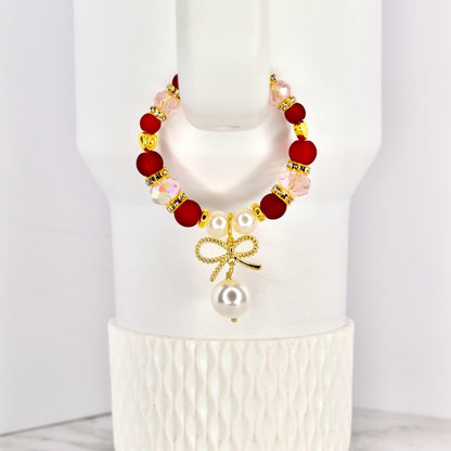A beaded tumbler handle bracelet with red, pink, and gold accents. The bracelet features a gold bow charm with a dangling pearl. The bracelet is displayed on a white Stanley.